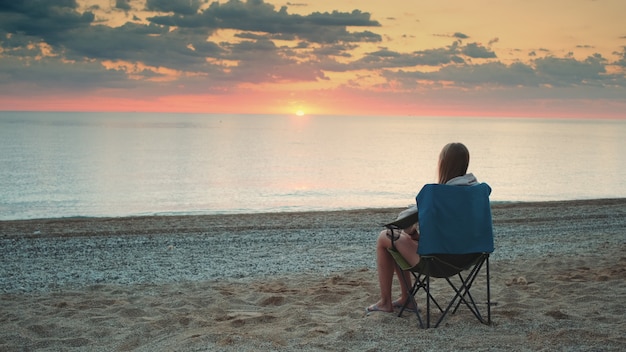 Mulher admirando o pôr do sol no mar, sentada em uma cadeira dobrável de turista