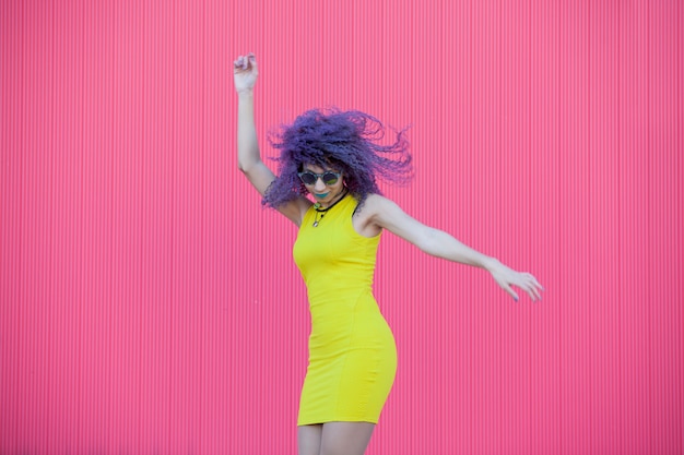 Mulher adolescente com óculos e cabelo afro roxo dançando em uma parede rosa