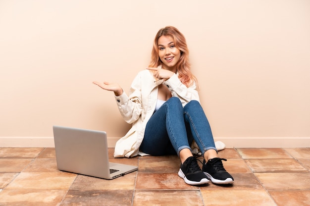 Mulher adolescente com um laptop sentada no chão em um ambiente fechado, segurando o imaginário de copyspace na palma da mão para inserir um anúncio