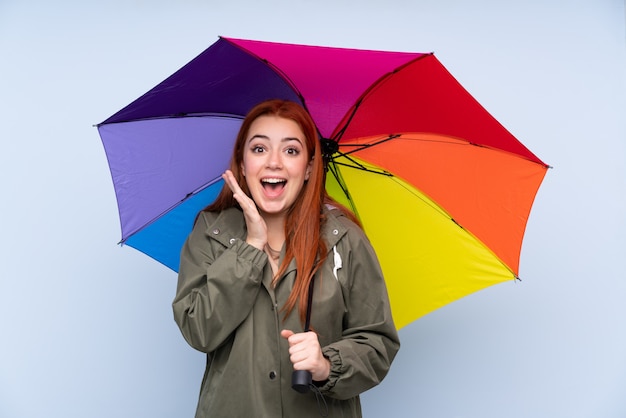 Mulher adolescente ruiva segurando um guarda-chuva sobre parede azul isolada com expressão facial de surpresa