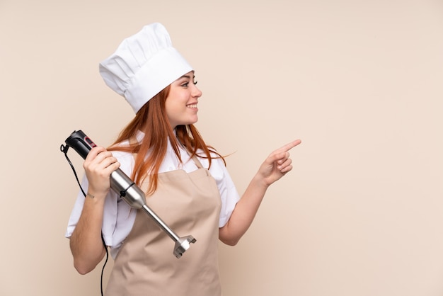 Mulher adolescente ruiva usando liquidificador mão sobre parede isolada, apontando para o lado para apresentar um produto