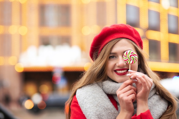 Mulher adorável e alegre vestida com um casaco vermelho e um lenço quente segurando um doce de caramelo na rua no inverno