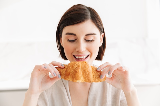 Foto mulher adorável e com fome 20 anos de roupão tomando café da manhã no apartamento, enquanto come croissant saboroso com prazer