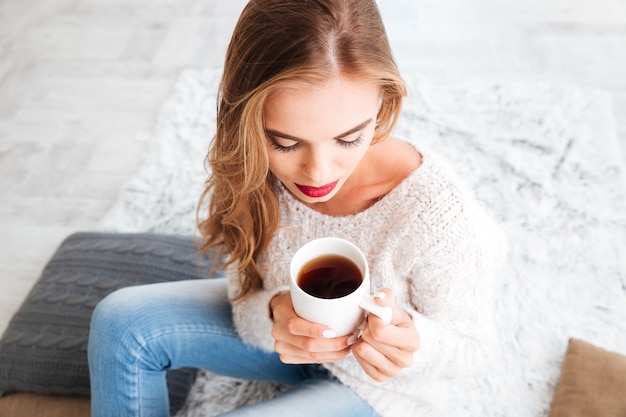 Mulher adorável e fofa com cabelo comprido e batom vermelho segurando uma xícara de chá dentro de casa