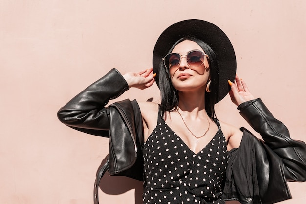 Mulher adorável elegante chapéu com jaqueta vestido com óculos de sol gosta de sol na cidade. Modelo de garota atraente moda em preto elegante verão lindas roupas posando sob a luz do sol perto da parede rosa