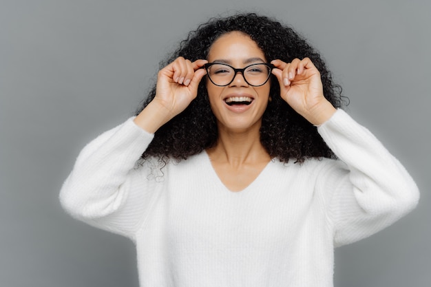 Foto mulher adorável otimista olha alegremente através dos óculos, mantém as mãos na borda dos óculos