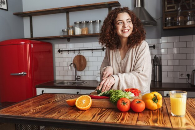 Mulher adorável sorrindo enquanto cozinha salada com legumes frescos no interior da cozinha em casa