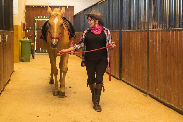 Mulher adulta caucasiana em um estábulo andando com um cavalo marrom, vestida com roupas da América do Sul e trabalhando com cavalos