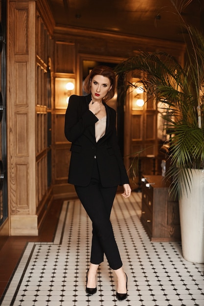 Mulher adulta elegante com maquiagem de noite e lábios vermelhos, vestindo um elegante terno preto posando no interior vintage