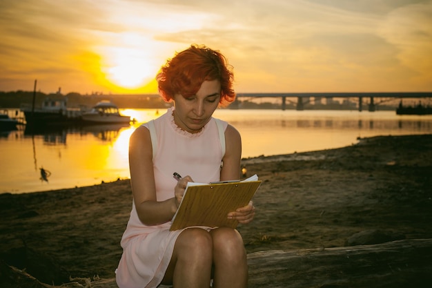Foto mulher adulta em meio a escrever sentada na margem do rio contra o céu durante o pôr-do-sol