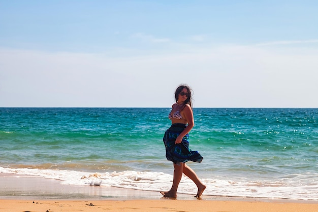 Mulher adulta em roupas de praia azuis e óculos de sol andando olhando para longe no mar azul