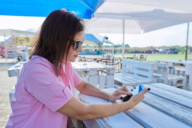 Mulher adulta em um café ao ar livre vazio em uma cidade deserta usando um smartphone,