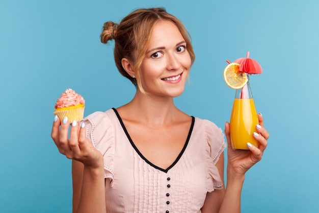 Mulher adulta jovem com aparência agradável, segurando nas mãos um delicioso bolo e coquetel de frutas, olhando para a câmera com expressão facial feliz. Tiro de estúdio interior isolado sobre fundo azul.