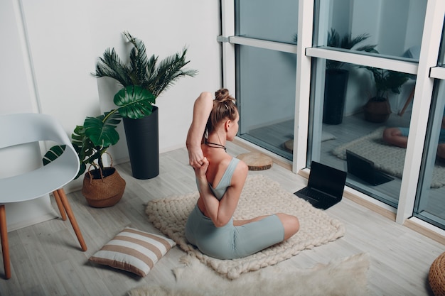 Mulher adulta madura fazendo ioga na sala de estar de casa com tutoriais on-line no laptop