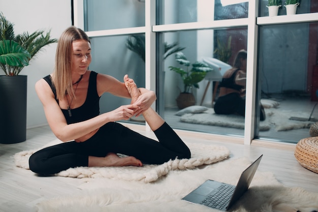 Mulher adulta madura fazendo ioga na sala de estar de casa com tutoriais on-line no laptop