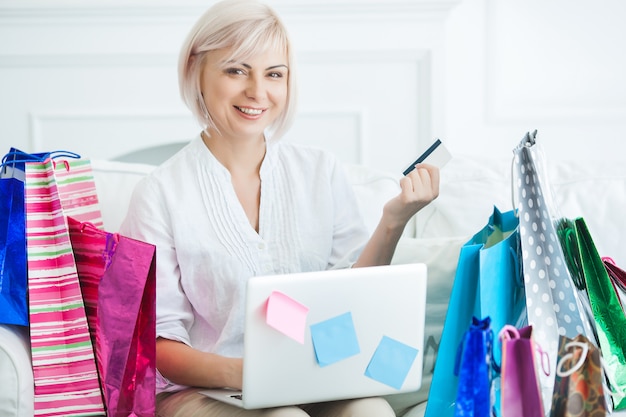 Mulher adulta meada atraente fazendo compras on-line. Cartão de crédito de exploração feminina e sentado no laptop com sacolas de compras dentro de casa.