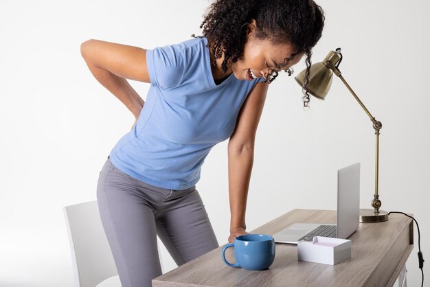 Foto mulher adulta média afro-americana com dor nas costas em pé ao lado do laptop, copo e lâmpada na mesa, espaço de cópia. fundo branco, tecnologia sem fio, dor nas costas, parte inferior, coluna vertebral, médico, dor, cuidados de saúde.