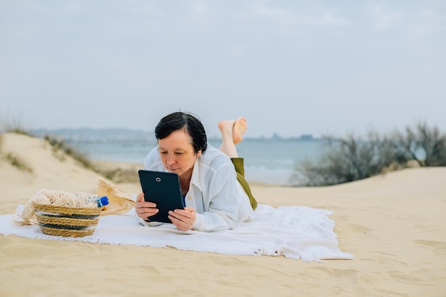 Mulher adulta na praia à beira-mar na primavera senta-se em um piquenique lê um livro em um tablet assiste a