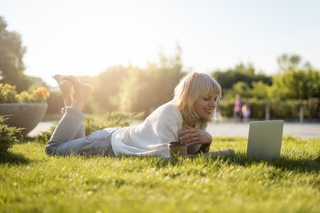 Mulher adulta olha para o laptop, deitado na grama do lado de fora no parque.