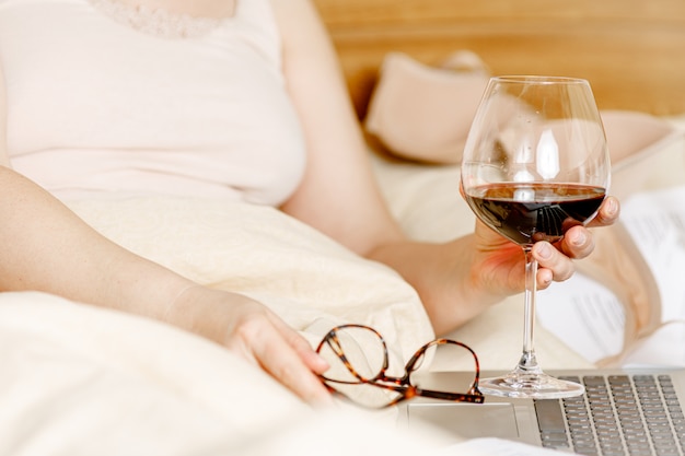 Mulher adulta relaxante na cama com um copo de vinho tinto e o computador portátil