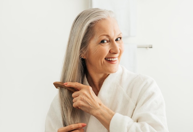 Mulher adulta sênior penteando o cabelo com um pente de madeira Mulher sorridente em um roupão de banho cuidando de seus longos cabelos grisalhosx9xA