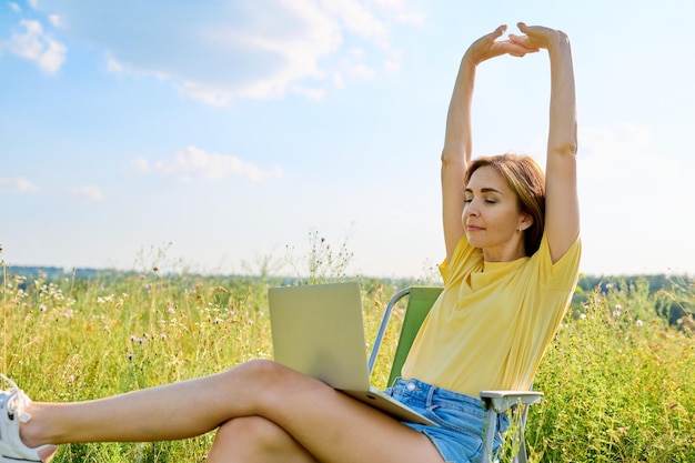 Mulher adulta sentada em uma cadeira de acampamento dobrável usando um laptop em um prado com ervas selvagens