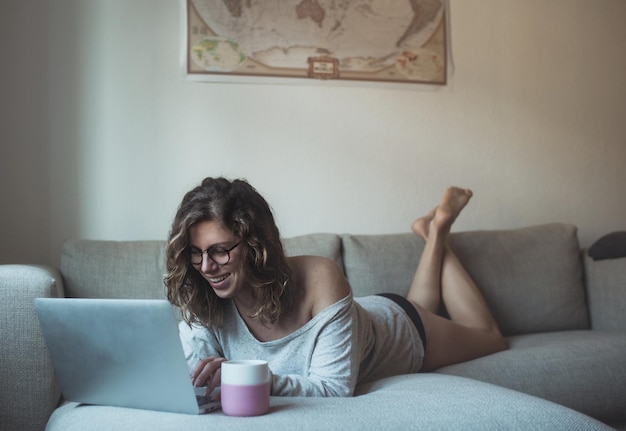 Mulher adulta sorridente usando laptop enquanto deitada no sofá em casa