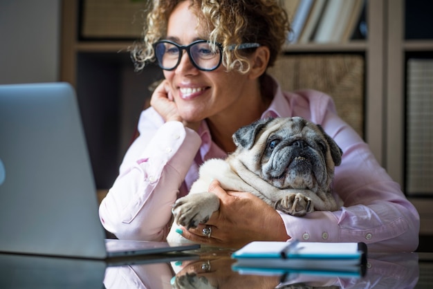 Mulher adulta trabalhando em um laptop em casa, no escritório, com seu adorável melhor amigo, cachorro pug