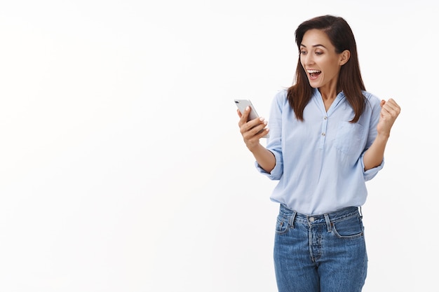 Mulher adulta triunfante alegre dos anos 30 sacode o punho enquanto lê excelentes notícias, sorri amplamente segura o smartphone, olha a tela do telefone móvel regozijando-se, ganha na loteria online, fica de pé na parede branca