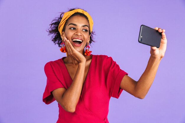 Mulher africana alegre num vestido fazendo selfie no smartphone enquanto toca sua bochecha sobre a parede roxa