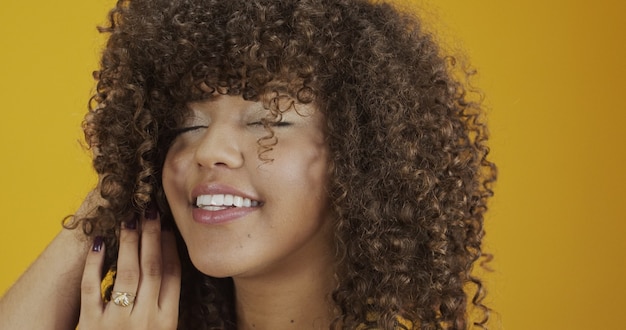 Mulher africana americana rindo feliz com seu cabelo encaracolado em fundo amarelo. Rindo mulher encaracolada de suéter, tocando seu cabelo e olhando para a câmera.