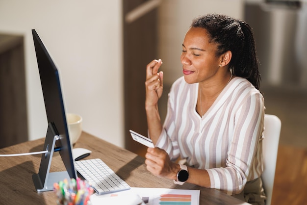 Mulher africana antecipando com os olhos fechados e os dedos cruzados esperando boas notícias enquanto usa cartão de crédito e trabalha no computador.