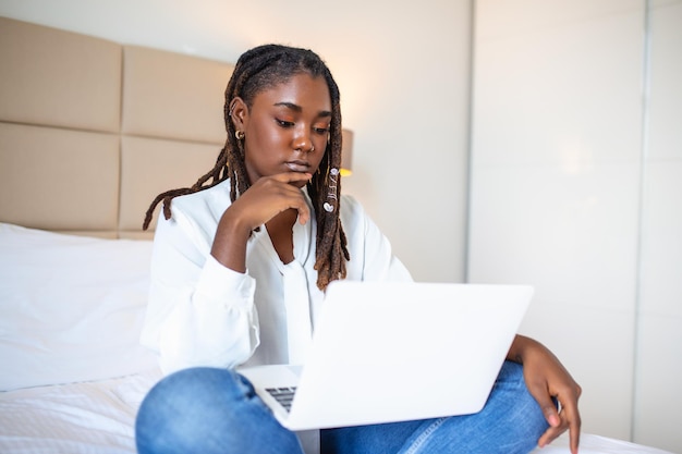 Mulher africana atraente de camisa branca usando laptop e sorrindo pela manhã Chat de vídeo e freelance trabalhando em casa conceito de bela jovem com laptop na cama