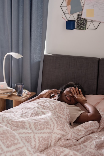 Mulher africana cansada deitada na cama e falando no celular no quarto dela