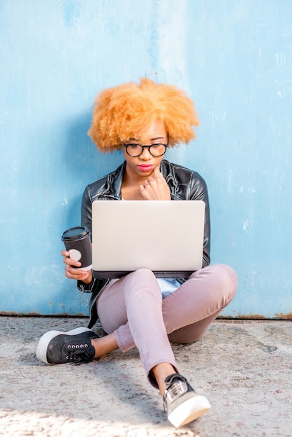 Mulher africana com cabelo encaracolado trabalhando com laptop sentada no fundo da parede azul