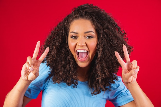 Mulher africana com seu cabelo encaracolado sobre fundo vermelho. Sinal de vitória. V. Rindo mulher encaracolada em olhar para a câmera.