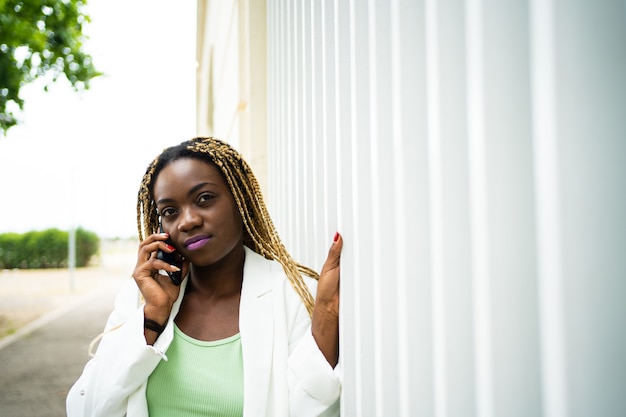 Mulher africana com tranças e roupas formais conversando com o celular ao ar livre