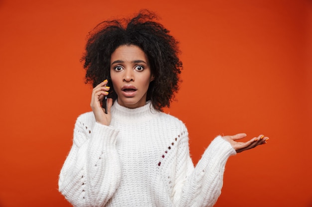 Mulher africana confusa e atraente usando um suéter falando no celular, isolado sobre a parede vermelha