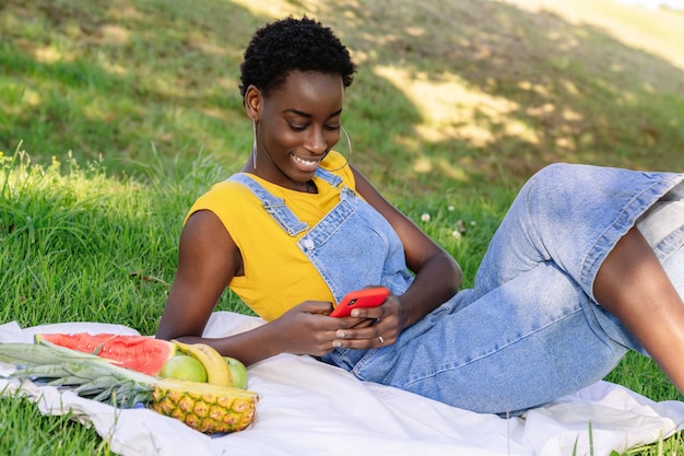 Mulher africana deitada em um parque ao ar livre conversando e enviando mensagens de texto em seu celular enquanto fazia um piquenique