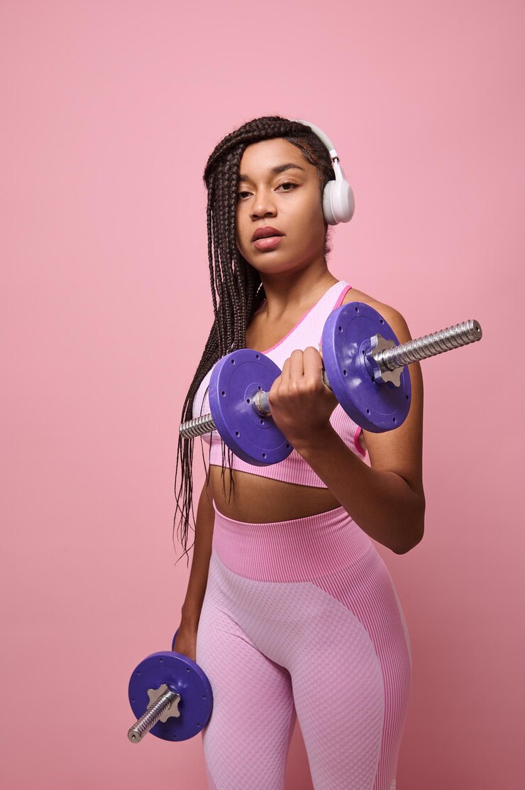 Foto mulher africana determinada com fones de ouvido sem fio, vestindo roupas esportivas rosa, fazendo exercícios no bíceps, isolado sobre um fundo rosa com espaço de cópia para anúncio
