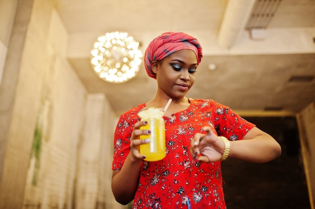 Mulher africana elegante de camisa vermelha e chapéu posou café interior e beber limonada de abacaxi