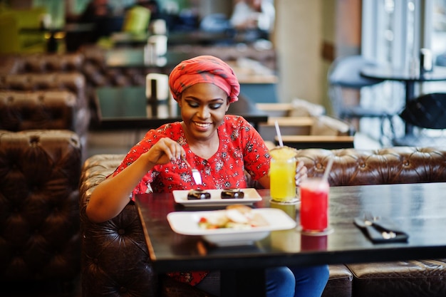 Mulher africana elegante de camisa vermelha e chapéu posou café interno come bolos de sobremesa de chocolate