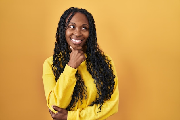 Mulher africana em pé sobre fundo amarelo olhando confiante para a câmera sorrindo com braços cruzados e mão levantada no queixo. pensando positivo.