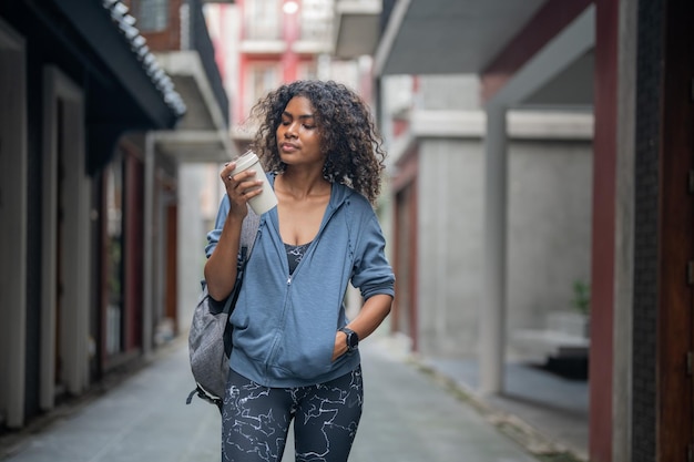 Mulher africana esporte ajuste saudável e magro firme bebe água do copo na mão garota fitness vestindo roupas esportivas e jaqueta andando na cidade e cidade Fitness e motivação esportiva