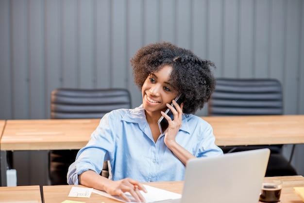 Mulher africana falando com o telefone sentada com um laptop e documentos no interior do escritório moderno