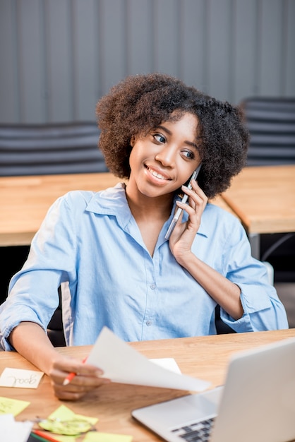 Mulher africana falando com o telefone sentada com um laptop e documentos no interior do escritório moderno