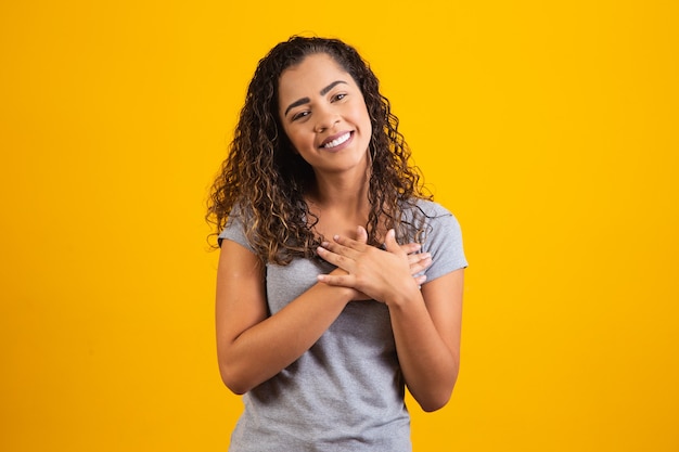 Foto mulher africana feliz em pose de camiseta cinza em fundo amarelo, sorrindo, olhando para a câmera de mãos dadas no peito, sentindo gratidão, coração, gesto de sentimentos sinceros e conceito de amor