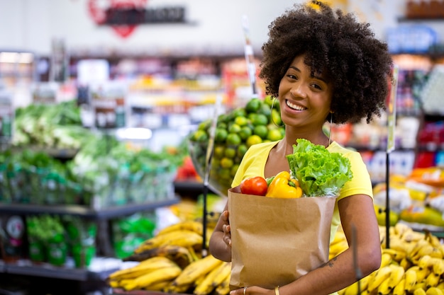 Mulher africana feliz positiva saudável segurando uma sacola de papel cheia de frutas e legumes.