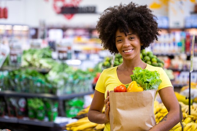 Mulher africana feliz positiva saudável segurando uma sacola de papel cheia de frutas e legumes.