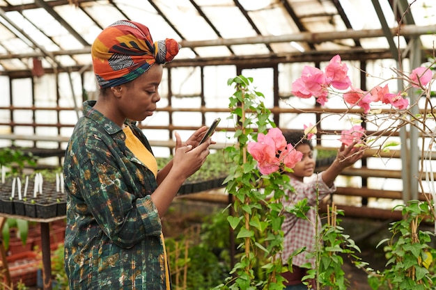 Mulher africana fotografando flores em seu telefone celular enquanto cultiva plantas em uma estufa